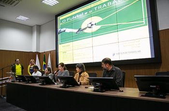 foto da mesa de debates com os palestrantes sentados e ao fundo arte do evento no telão