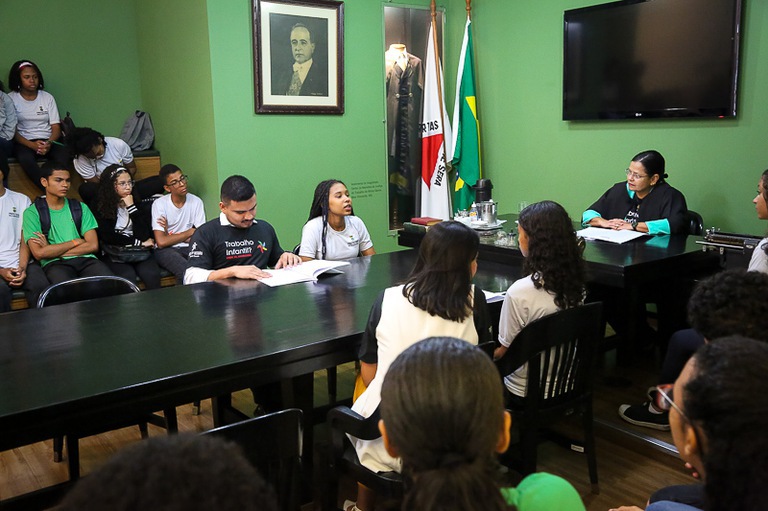 Adolescentes sentados em uma mesa de madeira observam desembargadora explicar o funcionamento de uma audiência trabalhista.