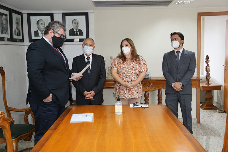 Foto do momento em que o Diretor-Geral do TRT-MG lê o termo de posse e o presidente, a empossanda e o juiz auxiliar da presidência prestam atenção. Ao centro há uma mesa grande e, ao fundo, há um aparador e fotos de alguns ex-presidentes do Tribubal penduradas na parede.