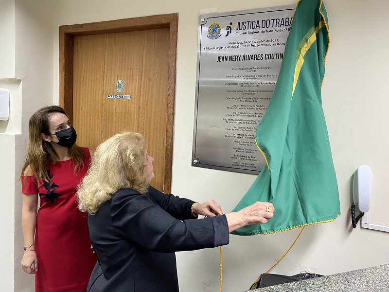 Foto da viúva do homenageado, Ondina Maria de Souza Coutinho, descerra a placa ao lado da juíza Martha Halfeld. A viúva étem cabelos loiros no ombro e veste um casaco escuro. Ajuiíza tem cabelos longos castanhos e usa um vestido vermelho.