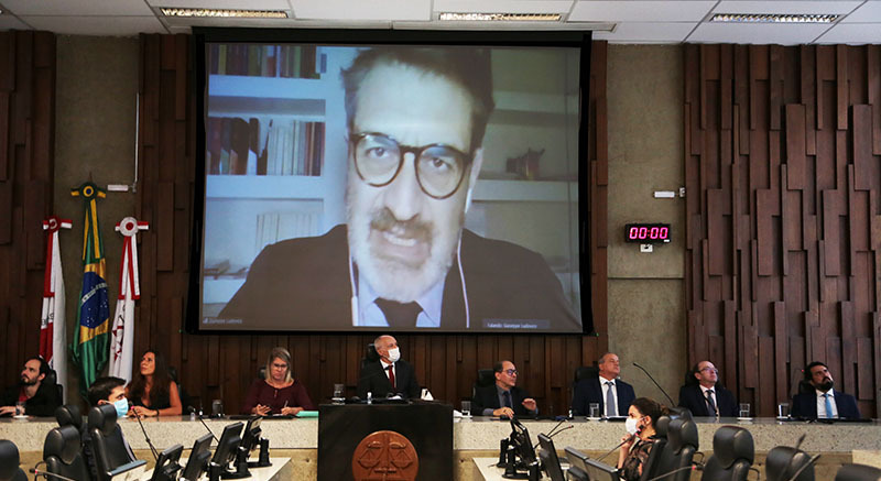 Participantes do evento sentados lado a lado na mesa com a imagem do professor italiano, um homem branco, sendo exibida atrás deles em telão