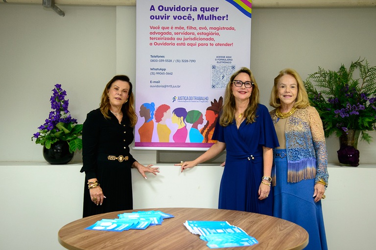 foto das desembargadoras Paula Cantelli, Emília Fachini e Maria Cristina Caixeta na frente de banner da Ouvidoria da Mulher