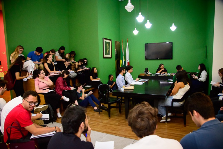 Alunos assentados assistem a uma audiência simulada. Os participantes da audiência estão assentados frente a frente em uma mesa preta retangular. Na cabeceira na mesa, está a desembargadora. 