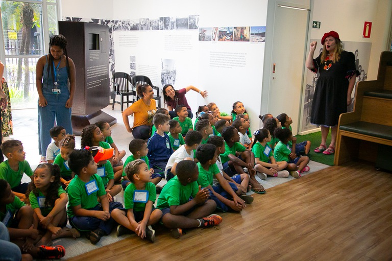 foto da contadora de histórias em pé falando para os alunos de uniforme verde sentados