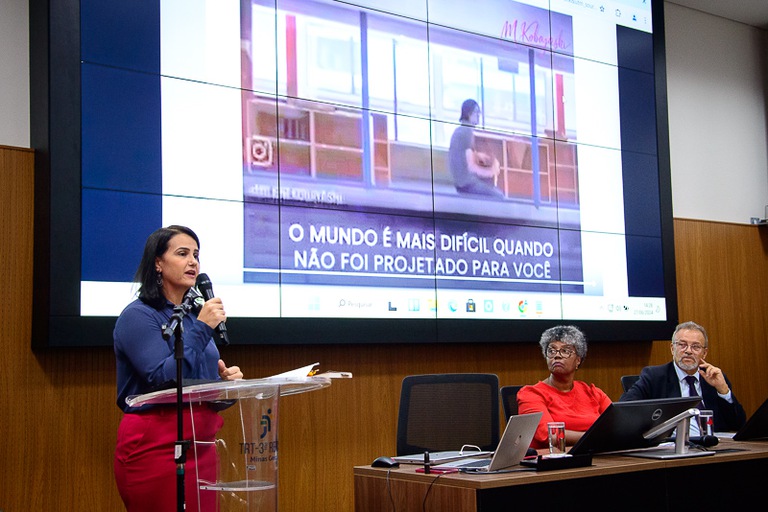 foto da palestrante juíza do TRT-MG, Jéssica Grazielle Andrade Martins, em pé com a mesa de honra ao lado 