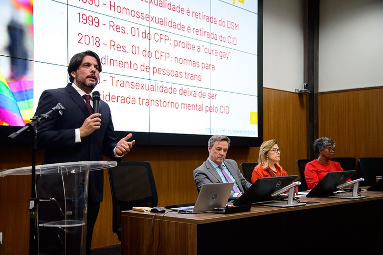 foto do palestrante professor da UFOP, Alexandre Melo Franco Bahia, em pé falando, ao lado da mesa de honra