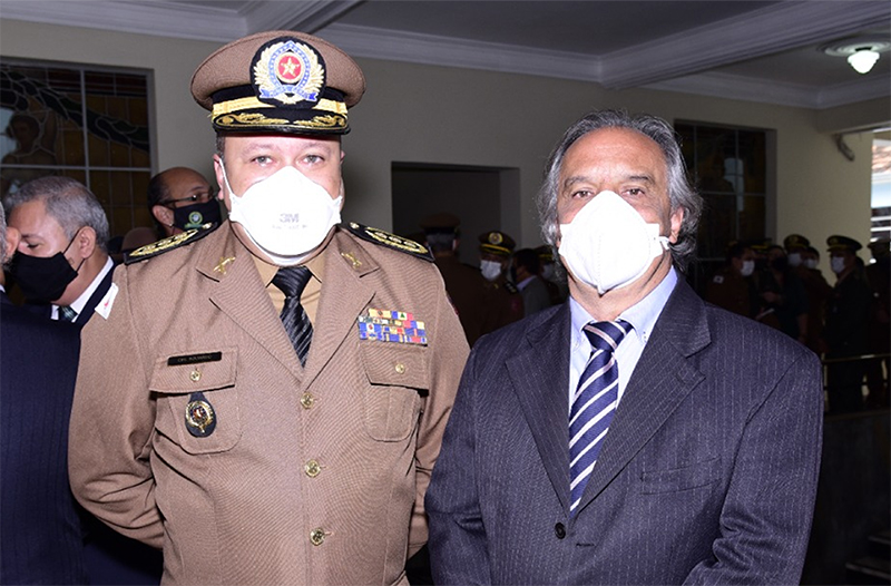 - Desembargador Paulo Maurício e o coronel Eduardo Felisberto Alves, chefe do Estado Maior da PMMG. Os dois usam máscaras e o coreonal está uniformizados. Ao fundo há vários convidados do evento formando grupos de conversa. 