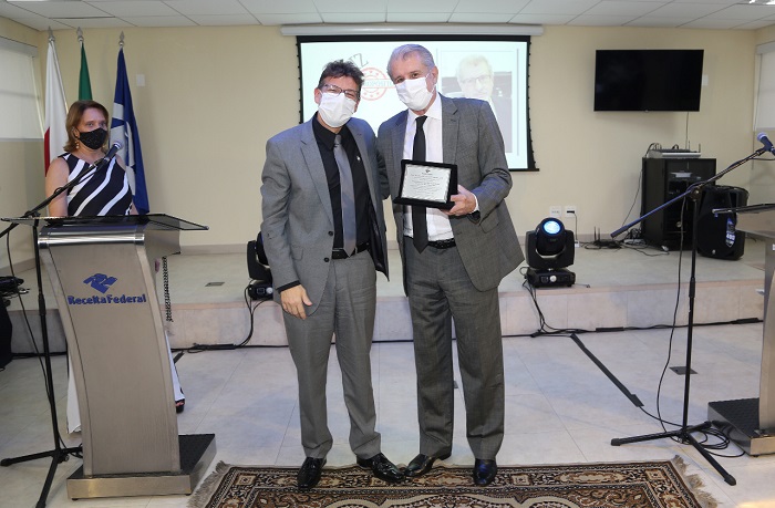 Foto do Superintendente da RFB em MG ao lado do presidente do TRT-MG, que segura a plca com a homenagem que recebeu. À esquerda, a mestre de cerimônia atrás de um púlpito e, ao fundo, um telão. 
