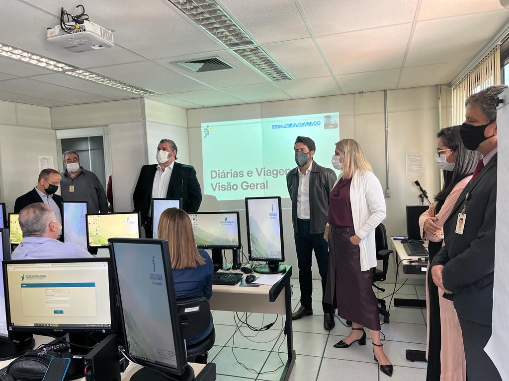 Foto com algumas pessoas de pé formando uma meia lua. Eles estão de frente para  outras pessoas assentadas de frente para telas de  computadores
