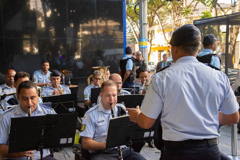 Banda de Música da Guarda Civil Municipal de Belo Horizonte se apresentando de farda e sentados tocando instrumentos