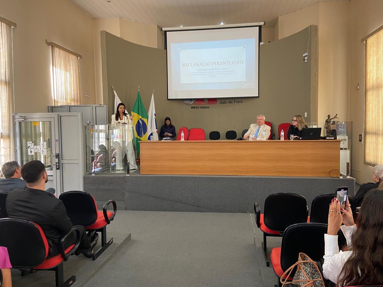 Palestrantes em pé e sentados perto da mesa de honra com a plateia na frente 