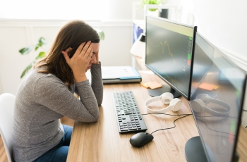 Foto de uma mulher em sua mesa de trabalho em frente ao computador, com as mãos no rosto e na testa. Ela é vista em posição de perfil com uma expressão de preocupação e cansaço.