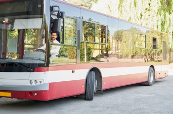 Imagem de um motorista de ônibus fazendo manobras no veículo vermelho e branco. A roda dianteira do ônibus está virada para a esquerda.