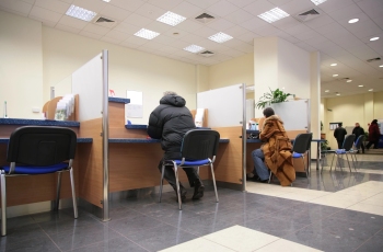 Na imagem, aparecem algumas pessoas de costas no interior de uma pequena agência bancária. Elas estão sentadas em cadeiras de escritório e sendo atendidas em vários guichês da agência bancária bem iluminada.