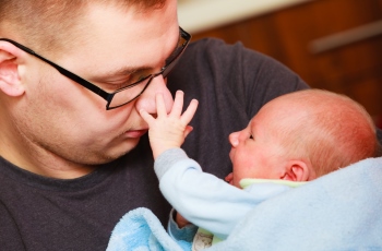 Foto de um pai interagindo com o filho recém-nascido. O pai carrega o filho no colo com um olhar de ternura, enquanto o bebê dá risada e se diverte puxando o nariz do pai.