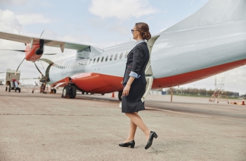 Foto de uma mulher caminhando entre aviões na pista de um aeroporto. Ela está usando blazer e saia preta e um sapato de salto alto elegante.