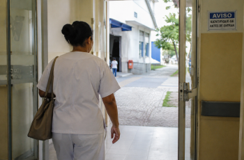 Imagem de um corredor de hospital brasileiro bem iluminado. Em primeiro plano, uma mulher adulta vista de costas usando uniforme hospitalar e segurando uma bolsa. Ela caminha em direção à saída, enquanto o ambiente hospitalar permanece ao fundo.