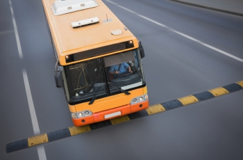 Foto de um ônibus, em péssimo estado de conservação, lotado de passageiros, passando em alta velocidade em cima de um quebra-molas. O motorista pode ser visto através da janela de vidro do ônibus.