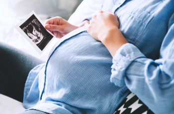 Foto de uma mulher grávida sentada em um sofá confortável. Ela está acariciando a barriga com uma das mãos e, com a outra, ela está segurando um exame impresso de ultrassom. Não aparece o rosto da gestante.