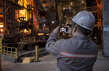 Foto de um homem metalúrgico uniformizado fotografando com um smartphone o am-biente ao redor dentro da usina siderúrgica onde ele trabalha.
