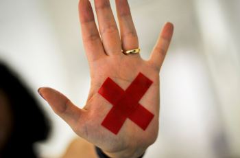 Foto de mão feminina que contém o desenho de um X vermelho, símbolo da luta contra a violência doméstica e familiar contra a mulher.