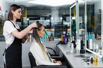 Foto de uma mulher usando um avental preto, exercendo a função de cabeleireira. Ela está fazendo os últimos retoques nas mechas com tintura dourada de uma mulher negra. Ela está terminando de fazer um penteado de tranças afro na cliente, que está sentada na cadeira do salão de beleza, cercada de cosméticos e espelhos grandes.
