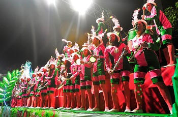 Foto de pessoas alegres cantando fantasiadas no Carnaval tocando pandeiro e outros instrumentos musicais.