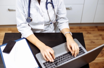 Foto de uma médica usando um estetoscópio e digitando no notebook para atender consultas on-line. Ao lado dela, na mesa de trabalho, há um celular e um receituário médico. Não aparece o rosto dela.