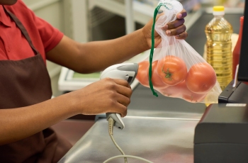 Na imagem, aparecem as mãos de uma operadora de caixa no supermercado segurando uma embalagem plástica com tomates e uma máquina registradora de preços.