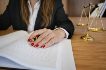 Foto de uma mulher com unhas pintadas de vermelho e cabelos compridos castanhos claros, contrastando com o casaco preto. Ela está com uma caneta na mão analisando documentos de um processo. Não aparece o rosto dela. Há uma balança dourada da justiça sobre a mesa de trabalho. Ao fundo, aparecem livros cuidadosamente organizados em uma estante.