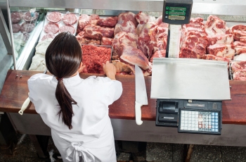 Foto de uma açougueira atendendo clientes no supermercado. Ela está pegando carne na gôndola do supermercado para pesar na balança que está ao lado dela.