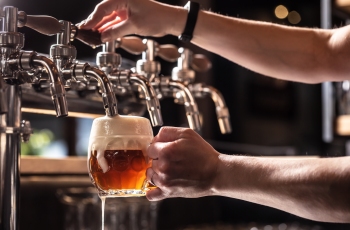 Foto do empregado da cervejaria enchendo uma caneca de vidro arredondada, com cerveja de espuma branca. Na foto só aparecem mãos e braços do homem.