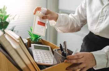 Imagem de uma mulher organizando os seus pertences em uma caixa, após ser dispensada do emprego. Há um crachá, calculadora e canetas dentro caixa, entre outros objetos. Não aparece o rosto da mulher.