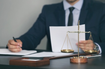 Foto de homem de terno e gravata examinando documentos de um processo e fazendo anotações. Há uma balança da justiça sobre a mesa de trabalho dele.