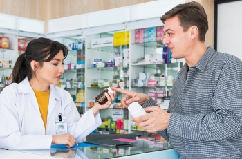 Foto de uma empregada de drogaria, que entrega frascos de medicamentos a um cliente que acabou de comprá-los. Ela usa jaleco e crachá e está no balcão da drogaria.