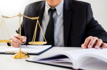 Foto de um homem de terno examinando documentos de um processo e fazendo anotações. Há uma balança símbolo da Justiça sobre a mesa de trabalho dele. Não aparece o rosto do homem.