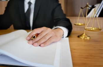 Foto de um homem usando terno e gravata. Ele está fazendo anotações e examinando os documentos de um processo. Há uma balança da justiça sobre a mesa de trabalho dele e alguns livros na estante ao fundo. Não aparece o rosto dele.