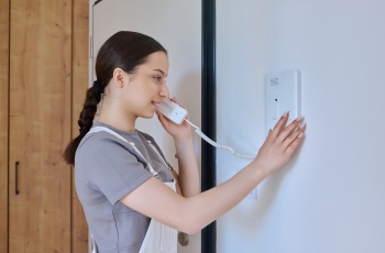 Foto de uma empregada doméstica, usando avental, atendendo a uma chamada pelo interfone.