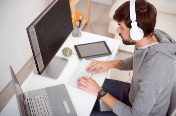 Visão lateral de um homem sentado trabalhando em frente ao computador. Ele está usando fones de ouvido.