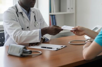 Foto de um médico negro usando jaleco branco e um estetoscópio em volta do pescoço. Ele entrega um atestado médico a um paciente de cabelos negros, que está usando uma camisa azul-turquesa. Há um receituário, caneta e um aparelho medidor de pressão arterial sobre a mesa de trabalho.