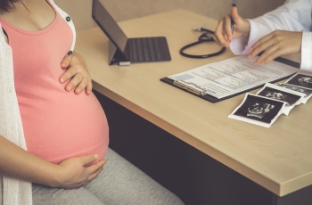 Foto de uma mulher grávida fazendo o pré-natal. Ela está com as mãos sobre a barriga, enquanto ouve as orientações do médico. Em cima da mesa de trabalho do médico há um estetoscópio, um laptop, uma prancheta com laudo médico e vários exames de ultrassom da paciente.