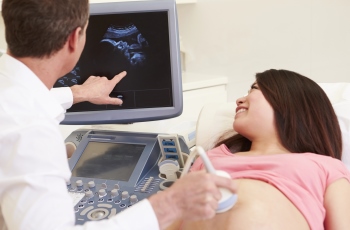 Foto de uma mulher grávida deitada sorrindo, fazendo o exame de ultrassom. Ao lado dela, o médico aponta para a tela que mostra a imagem do feto.