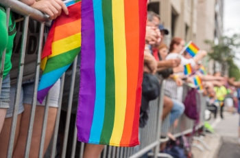Foto de várias pessoas segurando bandeira cor de arco-íris, símbolo do orgulho LGBTQIA+, enquanto assistem ao evento e também participam da parada LGBTQIA+ e das campanhas de combate à homofobia e transfobia.