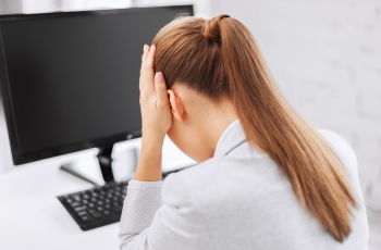 Foto de uma mulher de costas em frente à tela de um computador. Ela faz gestos com a mão no rosto que demonstram cansaço e preocupação. Ela usa o cabelo preso em rabo de cavalo.