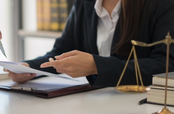 Foto de mulher usando blazer preto, examinando documentos de um processo. Há uma balança da justiça e livros empilhados sobre a mesa de trabalho dela.