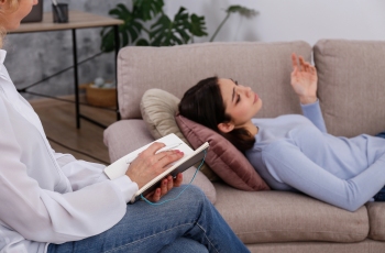 Foto de uma mulher deitada no divã fazendo psicanálise no consultório de uma psicoterapeuta, que está ouvindo atentamente e fazendo anotações.