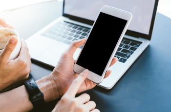 Foto de mãos femininas no ambiente de trabalho, ligando o smartphone para checar e analisar informações importantes. Ao lado, mãos de um colega de trabalho segurando uma xícara de café e, ao fundo, um notebook ligado.