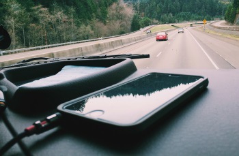 Foto de um smartphone conectado ao carregador de bateria, posicionado em cima do painel de um carro. O veículo está em movimento na rua.