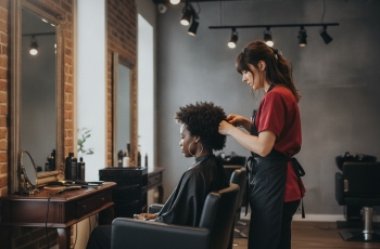 Foto de uma cabeleireira vista de perfil trabalhando em um salão de beleza comum, cuidando do cabelo black power de uma cliente negra que também é vista de perfil e está diante do espelho.