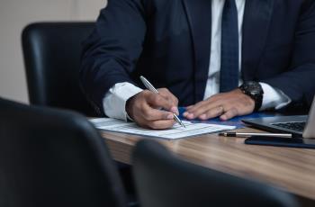Foto de um homem de terno e gravata examinando documentos de um processo e fazendo anotações. Há notebook e smartphone sobre a mesa de trabalho dele.